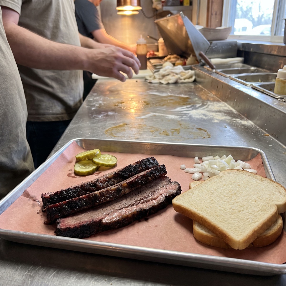 Slices of smoked brisket with a pink smoke ring on butcher paper, with pickles, chopped onion and white bread