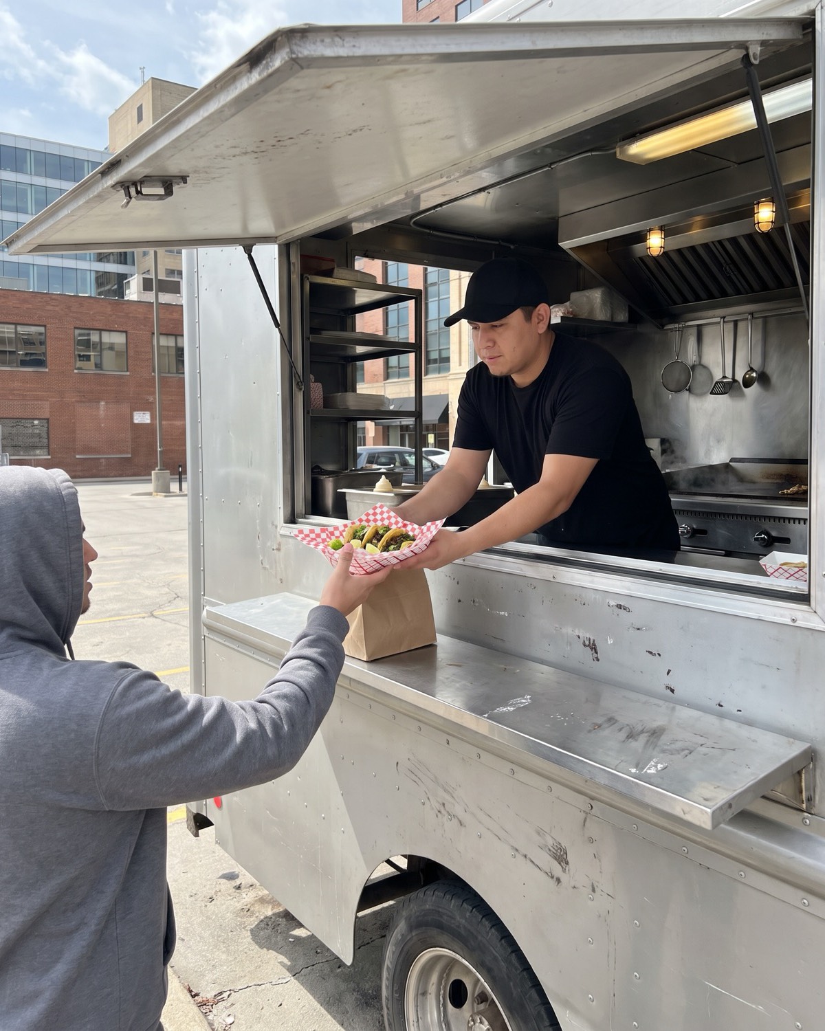 A cook hands a paper boat of tacos down from a food truck window to a customer at lunchtime